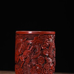 Puede incluir: Un jarrón de laca de cinabrio rojo con intrincados tallados de dragones y flores. El jarrón tiene una base redonda y un cuerpo cilíndrico.
