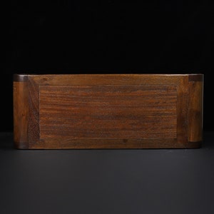 May include: A rectangular wooden box with a rich, dark brown finish. The box has a simple, elegant design with a flat top and rounded corners. The wood grain is visible, adding texture and warmth to the piece. The box is likely used for storage or display.