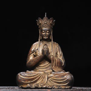 May include: A bronze-toned statue of a seated figure wearing an ornate crown with small relief carvings. The figure is in a meditative pose with hands pressed together in front of the chest, draped in flowing robes with visible folds and textures.