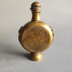 Puede incluir: Una pequeña botella redonda de latón con una tapa decorativa y tallas intrincadas. La botella tiene un diseño circular con caracteres y símbolos chinos.