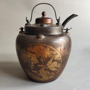 May include: A dark brown teapot with a gold design of birds and trees. The teapot has a metal handle and spout.