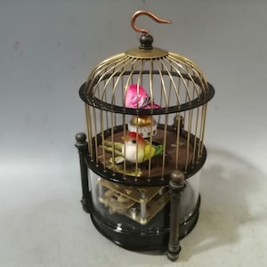 May include: A vintage birdcage music box with a glass base and two small, colorful birds inside. The cage is made of gold-colored metal and has a black base.