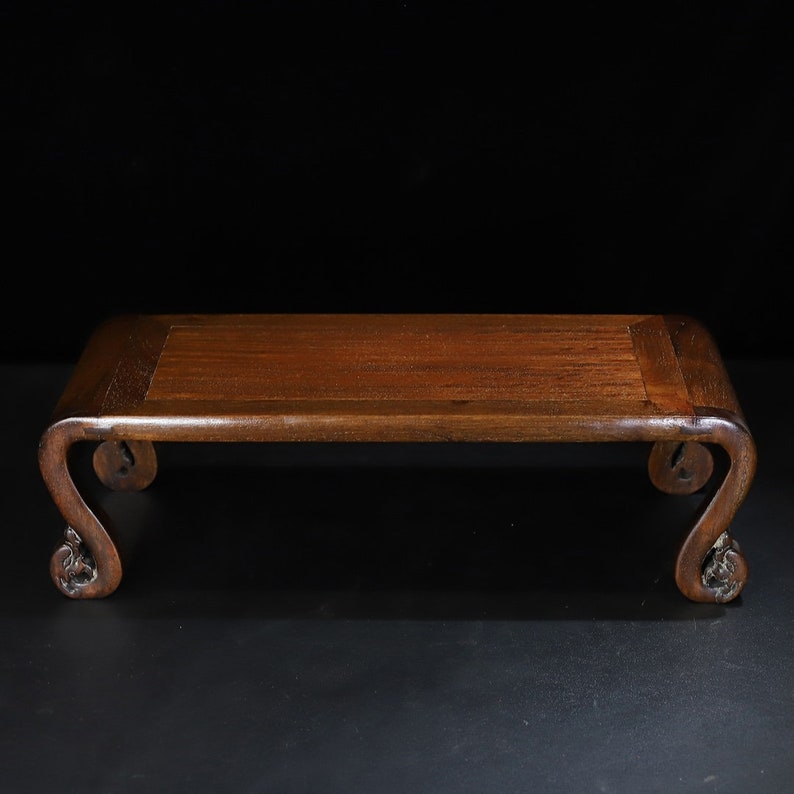 May include: A rectangular wooden display stand with a dark brown finish. The stand has curved legs with ornate carvings at the base. The top surface is smooth and rectangular, suitable for displaying small objects.