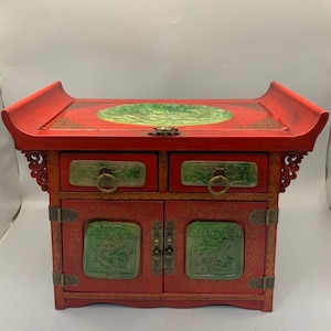 May include: A red wooden jewellery box with two drawers and a door. The box is decorated with green jade inlays and gold accents. The top of the box has a raised edge and a circular jade inlay.