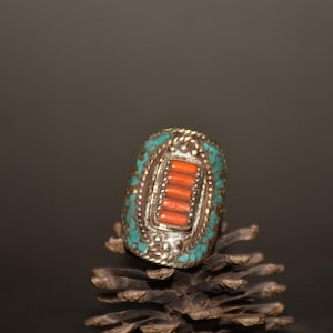 Puede incluir: Un anillo plateado con un diseño rectangular. El anillo presenta una fila central de piedras de coral naranja, rodeada de piedras de color turquesa y un borde decorativo. El anillo está sobre una piña.