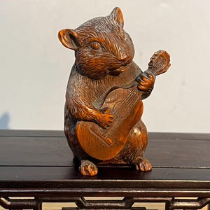 Puede incluir: Escultura de madera detallada de un roedor tocando un instrumento de cuerda. La escultura es de color marrón cálido, con vetas de madera visibles y detalles intrincados. El roedor está representado en posición sentada, sosteniendo el instrumento.