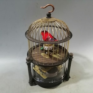 May include: A vintage, brass birdcage with a red and black bird inside. The cage has a glass base and a clock face on the bottom. The birdcage is on a white background.
