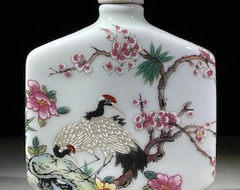 May include: White porcelain snuff bottle with a matching lid. The bottle is decorated with a painted scene of two cranes, pink cherry blossoms, and green leaves. The cranes have red crests and black and white feathers.