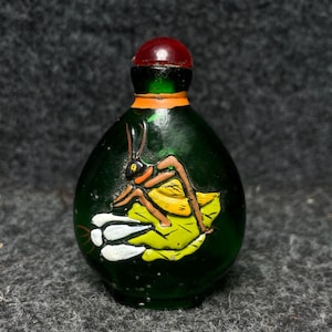 May include: A small, dark green glass bottle with a red stopper. The bottle features a painted insect design on a yellow leaf. The insect has brown legs and antennae. The bottle has an orange band around the neck.