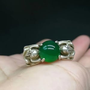 Puede incluir: Un anillo de plata con una gran gema verde vibrante. La banda del anillo tiene un diseño ornamentado con tallas detalladas. La gema es de forma ovalada y está engastada en un bisel de plata, creando un contraste llamativo.