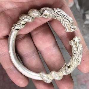 Pode incluir: Uma pulseira de prata com duas cabeças de dragão em cada extremidade. A pulseira é feita de um fio grosso e redondo, e os dragões são intrincadamente detalhados.