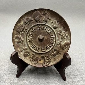 May include: A bronze-colored, circular antique plate featuring zodiac animal figures around the perimeter and Chinese characters in the center. The plate is displayed on a dark brown wooden stand.
