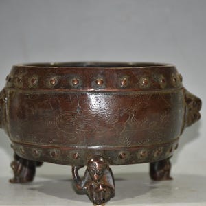 Può includere: Un bruciatore di incenso in bronzo con intagli intricati e manici a testa di leone. Il bruciatore ha tre gambe e un corpo rotondo.