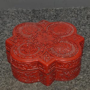 May include: A vibrant red, intricately carved box with a unique, multi-lobed shape. The box features detailed floral and geometric patterns across its surface, showcasing a high level of craftsmanship. The box is likely made of wood.