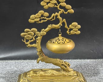 May include: A gold-toned metal incense burner featuring a sculpted tree branch base with detailed pine needle clusters. A rounded, perforated incense holder hangs from the branch by a small metal loop, all set upon a rectangular pedestal base.