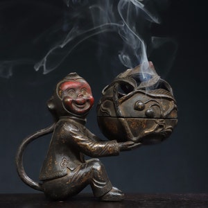 May include: A bronze-colored incense burner in the shape of a seated monkey holding a spherical container. The monkey has a red face and a smiling expression. Smoke rises from the container, suggesting the incense is lit. The container has a decorative leaf design.