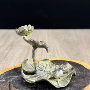 Könnte beinhalten: Ein metallener Räucherstäbchenhalter mit Antik-Finish, der ein geformtes Lotusblatt mit einem kleinen darauf ruhenden Frosch zeigt. Ein schlanker Stängel ragt aus dem Blatt hervor und trägt eine einzelne Lotusblütenknospe mit detaillierten Strukturen.