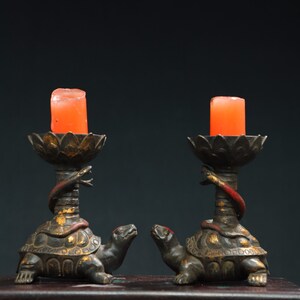 May include: Two black turtle candle holders with red candles. Each turtle has a gold snake wrapped around its neck and a gold lotus flower base.