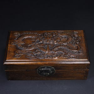 May include: A rectangular wooden box with a detailed carved dragon design on the lid. The box is a dark brown color and has a metal clasp on the front. The dragon is intricately carved, showcasing scales and flowing features. The box appears to be made of solid wood.