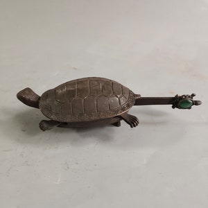 Puede incluir: Una pequeña figura de tortuga de bronce con una piedra preciosa verde en la cola. El caparazón de la tortuga está decorado con un patrón geométrico.