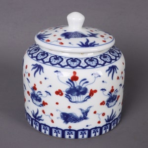Könnte beinhalten: Eine weiße Keramikdose mit Deckel, verziert mit blauen und roten Blumen- und Fischmotiven. Die Dose hat einen traditionellen chinesischen Stil.