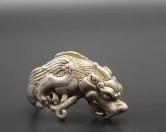 May include: A silver ring featuring a detailed, three-dimensional design of a mythical creature with wings. The creature's head is turned to the side, and its mouth is open, revealing sharp teeth.