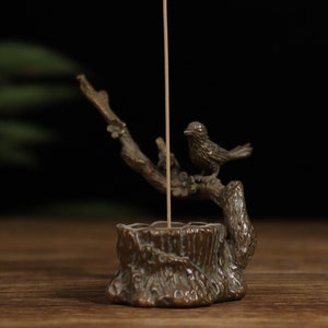 Puede incluir: Un quemador de incienso de bronce con forma de rama de árbol con un pájaro posado en él. El quemador de incienso tiene un pequeño agujero en el centro para colocar un palo de incienso.
