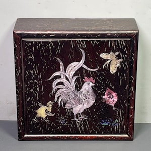 Puede incluir: Una caja de madera oscura con una incrustación decorativa que presenta un gallo, un pollito y mariposas. El gallo tiene un cuerpo blanco y rosa con una cresta roja. El pollito es amarillo y las mariposas son iridiscentes.
