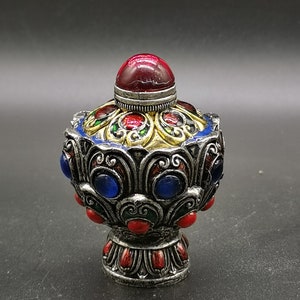 Puede incluir: Botella decorativa plateada ornamentada con un tapón rojo en forma de joya. La botella presenta detalles de joyas azules y rojas, con detalles dorados y verdes. El diseño es intrincado y detallado, lo que sugiere un estilo vintage o antiguo.