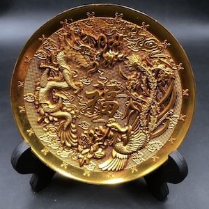Op de afbeelding: Een goudkleurige decoratieve plaat met een gedetailleerd reliëf van een draak en feniks, met Chinese karakters in het midden. De plaat wordt gepresenteerd op een zwarte standaard, die het ingewikkelde ontwerp benadrukt.