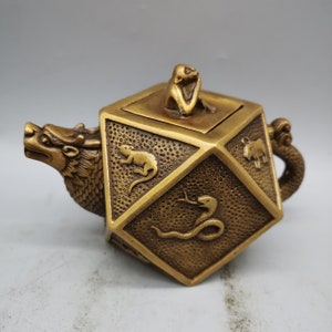 Puede incluir: Una tetera de latón con un pico de dragón y una tapa cuadrada con una figura de mono. La tetera está decorada con figuras de animales en relieve, incluyendo una rata, una serpiente y un buey.
