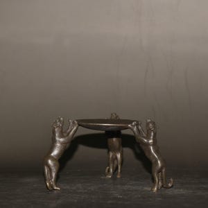 Könnte beinhalten: Bronze-farbener dekorativer Ständer mit drei Tierfiguren, die ein kreisförmiges Tablett tragen. Die Tiere sind in einer stehenden Pose dargestellt und halten das Tablett mit ihren Vorderpfoten. Der Ständer hat ein dunkles, gealtertes Aussehen.