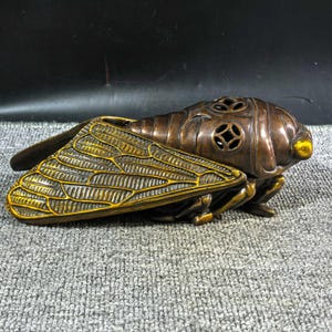 May include: A bronze-toned cicada sculpture with intricate wing and body detailing. The insect features a textured surface and decorative cutouts on its back. The sculpture is set against a grey background.