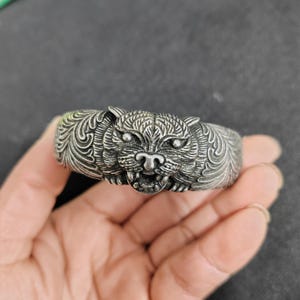 Könnte beinhalten: Ein silberfarbenes Armband mit einem detaillierten Tigerkopf-Design. Das Gesicht des Tigers ist aufwendig geschnitzt und zeigt Augen, Nase und geöffneten Mund. Das Armband hat eine strukturierte Oberfläche und einen klassischen, kühnen Stil.