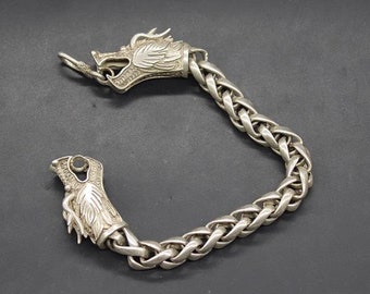 May include: Silver-toned bracelet featuring a dragon head design at each end. The bracelet has a thick, woven chain link pattern. The dragon heads have detailed scales and open mouths. The bracelet is displayed on a dark surface.
