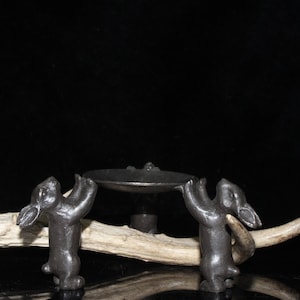 May include: A black metal incense burner with two rabbit figures holding up a round dish. The burner is on a wooden surface.