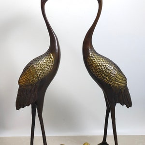 Op de afbeelding: Twee bronzen kraanvogelbeeldjes, elk staand op een schildpadbasis. De kraanvogels kijken elkaar aan, met hun kopjes omhoog gekanteld en hun snavels open. De beeldjes zijn gedetailleerd met gouden accenten.