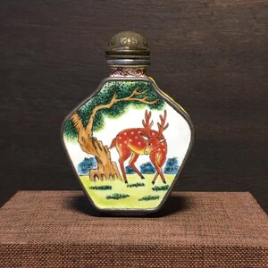 May include: A decorative, hand-painted glass snuff bottle with a metal lid. The bottle features a detailed scene of a deer under a tree, with a green and white background. The bottle has a silver-colored metal trim.