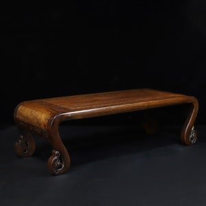 May include: A dark brown wooden display stand with a rectangular top and curved legs. The legs have a decorative, scroll-like design. The stand is made of a polished wood and is set against a black background.