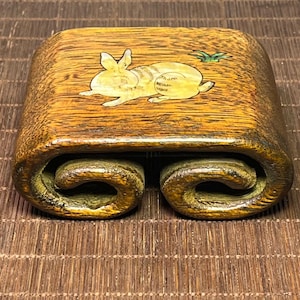 Puede incluir: Un soporte de rollo de madera con un diseño de conejo tallado. El soporte está pintado en tonos marrones y verdes.