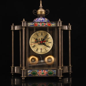 May include: A vintage-style brass clock with a glass case and a white face. The clock has a decorative top with blue and green enamel details. The clock face has Roman numerals and a black hour and minute hand. The clock is sitting on a black surface.