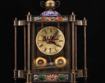 May include: A vintage-style brass clock with a glass case and a white face. The clock has a decorative top with blue and green enamel details. The clock face has Roman numerals and a black hour and minute hand. The clock is sitting on a black surface.