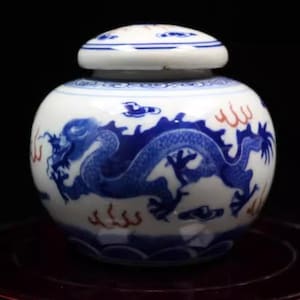 Op de afbeelding: Een kleine, witte porseleinen pot met een deksel, versierd met een blauw en rood drakenontwerp. De pot heeft een blauw en wit bloemenpatroon rond de rand.