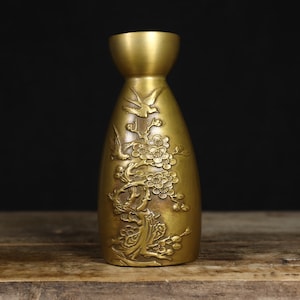 Peut inclure: Un vase en métal doré avec un col étroit et une base large. Le vase est décoré d'un motif en relief représentant une branche d'arbre fleurie avec des oiseaux.