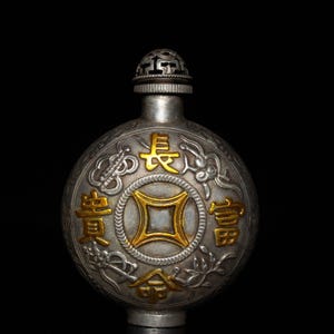 May include: A round, silver-toned metal flask featuring a central gold-colored square emblem surrounded by four gold Chinese characters. The surface is decorated with intricate embossed patterns and a filigree-style cap at the top of the vessel.
