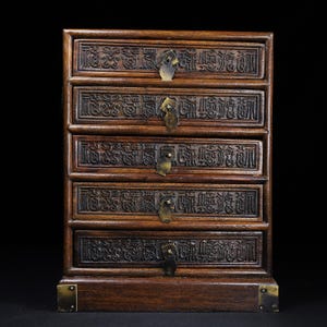May include: A dark brown wooden chest with five horizontal drawers. Each drawer front features intricate carved relief patterns and a small, centred brass pull handle. The base of the chest is reinforced with brass corner plates on the left and right.