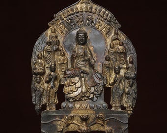 May include: A gold and black metal sculpture of a seated Buddha surrounded by smaller figures. The Buddha is in a meditative pose with hands in a mudra gesture. The sculpture is on a base with a lotus design.