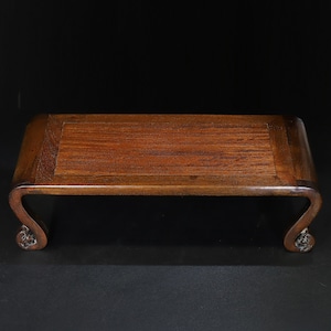 May include: A rectangular, wooden display stand with a dark brown finish. The stand features a flat top and curved legs with decorative carvings. The stand is designed to elevate and showcase objects.