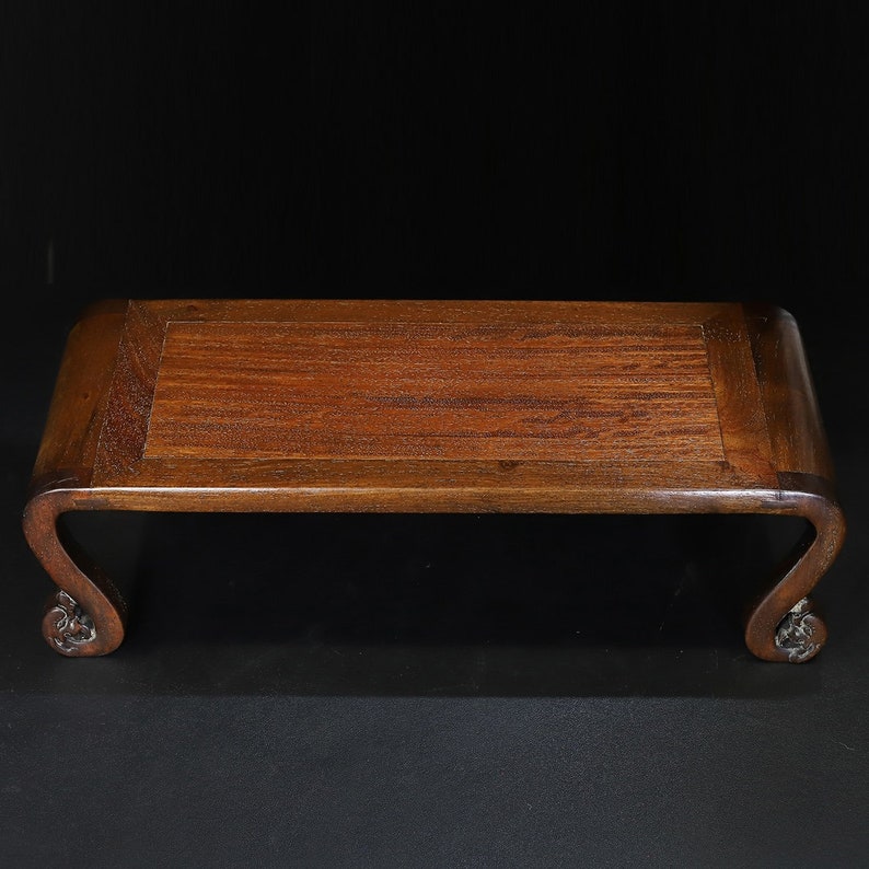 May include: A rectangular, wooden display stand with a dark brown finish. The stand features a flat top and curved legs with decorative carvings. The stand is designed to elevate and showcase objects.