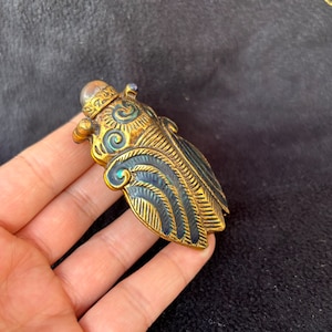 May include: A gold and blue enamel beetle-shaped pendant with intricate details. The pendant has a round, silver-coloured top with a small, clear stone.
